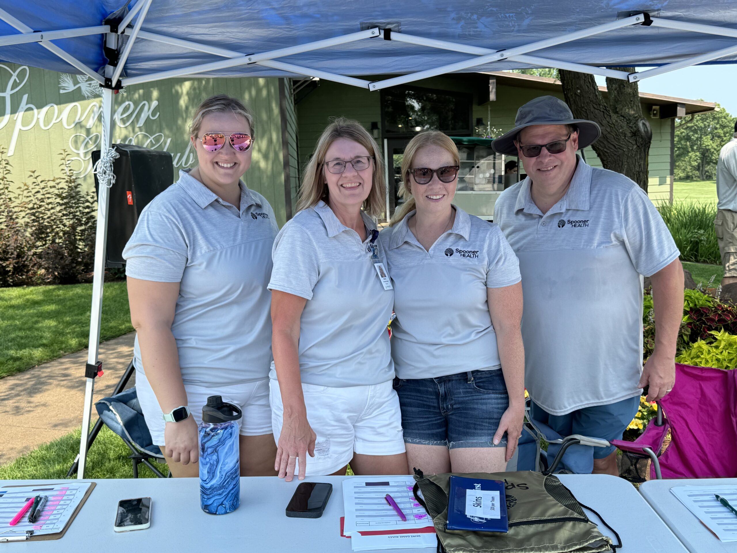 Spooner Health staff members at registration table during the Spooner Health Golf Outing, wearing matching shirts, smiling, with golf club in background.