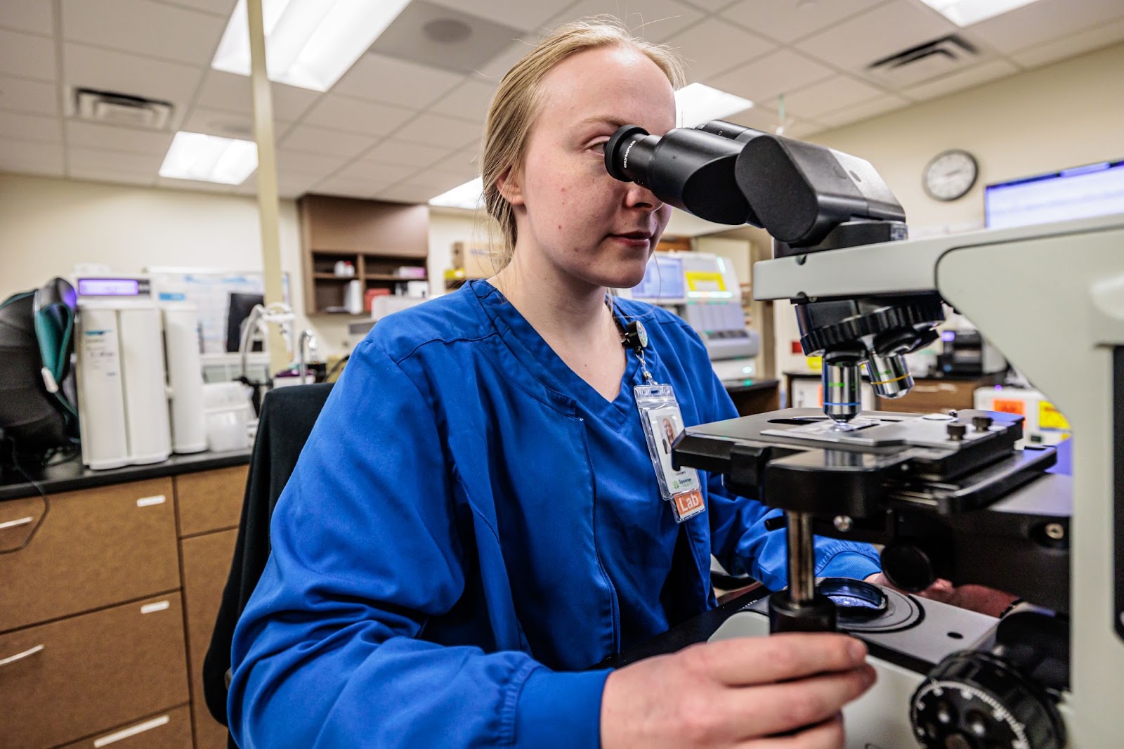 Laboratory technician in blue scrubs using a microscope for modern lab testing, emphasizing early disease detection at Spooner Health.