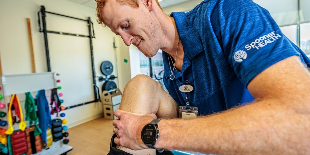 Physical therapist from Spooner Health assisting a patient with knee rehabilitation in a therapy setting, featuring exercise equipment and colorful resistance bands in the background.